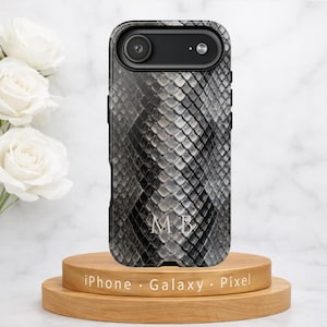 May include: A black and gray snake skin patterned phone case with the initials "MB" in white. The phone case is on a wooden stand with the words "iPhone · Galaxy · Pixel" printed on it. White roses are in the background.