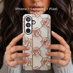 May include: A phone case featuring a stained glass-style pattern of pink roses on a cream background with gold lines. The text iPhone, Samsung, Pixel, and MagSafe is printed at the top. The case has a vertical camera cutout with three lenses.