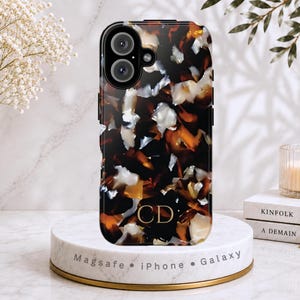 May include: A rectangular smartphone case featuring a glossy tortoiseshell pattern in shades of dark brown, amber, and white. The case includes a dual-lens camera cutout at the top and the gold-colored letters CD printed near the bottom center.
