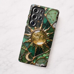 K&ouml;nnte beinhalten: Smartphone-H&uuml;lle mit einem Buntglas-Design, das ein goldenes Sonnengesicht, einen Mond und Sternbilder auf gr&uuml;nem Hintergrund zeigt. Die H&uuml;lle hat einen schwarzen Rand und einen Ausschnitt f&uuml;r mehrere Kameralinsen.