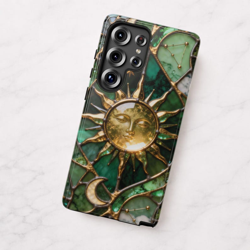 K&ouml;nnte beinhalten: Smartphone-H&uuml;lle mit einem Buntglas-Design, das ein goldenes Sonnengesicht, einen Mond und Sternbilder auf gr&uuml;nem Hintergrund zeigt. Die H&uuml;lle hat einen schwarzen Rand und einen Ausschnitt f&uuml;r mehrere Kameralinsen.