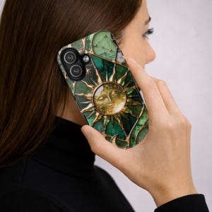 K&ouml;nnte beinhalten: Gr&uuml;ne und goldene Handyh&uuml;lle mit einem Sonnendesign. Die H&uuml;lle hat einen Glasmalerei-Effekt mit einem Sternbild-Design oben. Die Sonne hat ein Gesicht in der Mitte. Das Telefon wird ans Ohr gehalten.