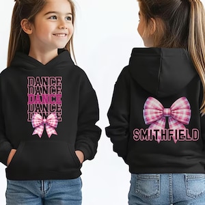 May include: Black hoodie with a front pocket, featuring the word "DANCE" stacked vertically with a pink plaid bow. The back of the hoodie has a matching bow and the name "SMITHFIELD" in pink.