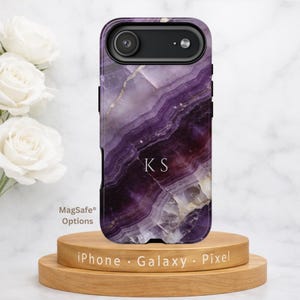 May include: A phone case with a purple and white marble design, featuring the initials "KS" in white. The case is displayed on a wooden stand with the text "iPhone · Galaxy · Pixel". The case has a black camera lens.
