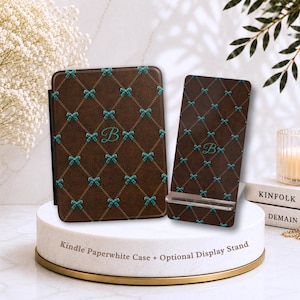 Pode incluir: Uma capa castanha para Kindle Paperwhite e um suporte a condizer, ambos com fundo castanho escuro, padrão de grelha de diamantes dourados, laços azul-petróleo nas interseções e um monograma com a letra B azul-petróleo ao centro.