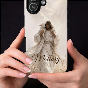 May include: A phone case featuring a printed illustration of a figure in flowing cream robes carrying a small white lamb on their shoulders. The design includes a field of wheat at the bottom and the name Mallory written in a brown cursive script.