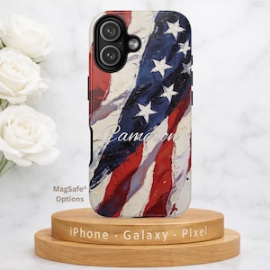 May include: A phone case with a painted American flag design. The case features red and white stripes, blue with white stars, and the name "Cameron" in cursive. The case is on a wooden stand with the text "iPhone Galaxy Pixel".
