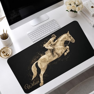 May include: A rectangular black desk mat featuring a gold-toned illustration of a rider on a jumping horse. The design includes a splattered paint effect and the name Wilson written in a cursive script at the bottom left corner of the mat.