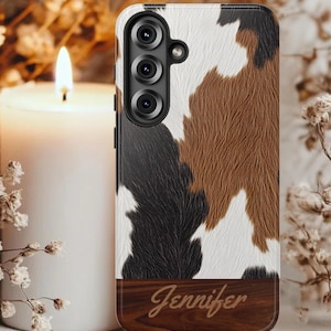 Könnte beinhalten: Smartphone-Hülle mit Kuhfellmuster in Schwarz, Braun und Weiß. Der untere Teil ist in dunkler Holzmaserung mit dem eingravierten Namen "Jennifer". Die Hülle hat ein Kameramodul mit drei Linsen.