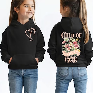 May include: A black hooded sweatshirt featuring a light pink heart and cross graphic with the name Natalie on the front. The back displays a floral bouquet graphic with the text Child of God and Psalm 23 in light pink lettering.