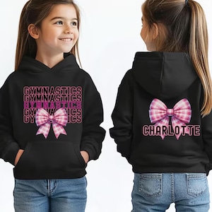 May include: A black hooded sweatshirt featuring the word GYMNASTICS repeated in pink and white text with a pink and white plaid bow graphic on the front, and a pink and white plaid bow with the name CHARLOTTE in pink text on the back.