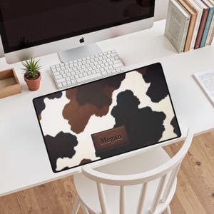 May include: A rectangular desk mat with a cowhide pattern in shades of brown, black, and white. A brown leather patch with the name "Megan" is centered at the bottom. The mat is on a white desk with a computer, keyboard, and other office supplies.