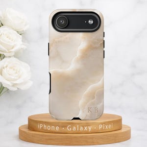 May include: A phone case with a cream-colored marble design, featuring black accents and a camera cutout. The case is displayed on a wooden stand with the text "iPhone Galaxy Pixel". The initials "KB" are visible on the case.