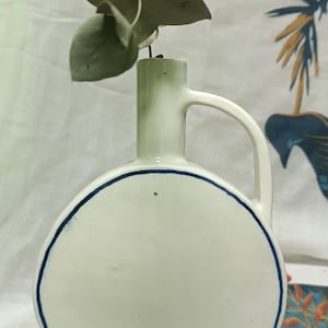 Puede incluir: Jarrón de cerámica blanca con un borde azul y un asa. El jarrón tiene un cuerpo redondo y un cuello estrecho. Una rama de eucalipto verde está en el jarrón.