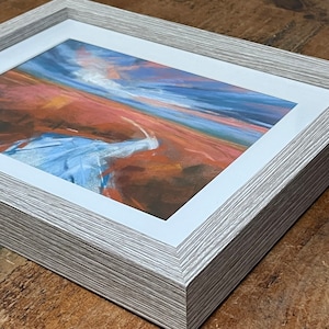 May include: A framed abstract landscape painting with a white mat and a light gray wood frame. The painting features a blue and white sky with a red and orange landscape.