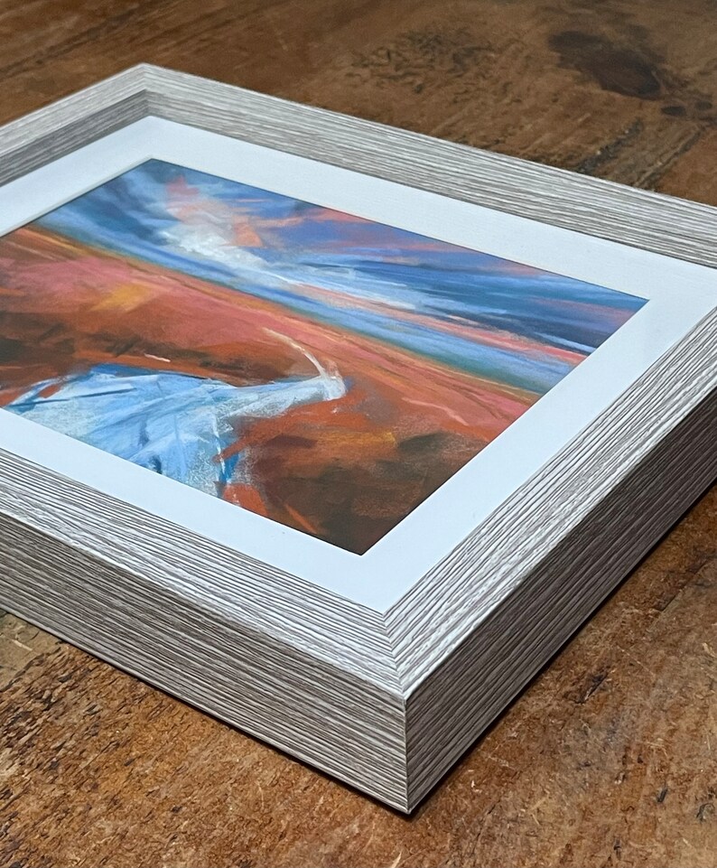 May include: A framed abstract landscape painting with a white mat and a light gray wood frame. The painting features a blue and white sky with a red and orange landscape.