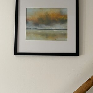May include: A framed painting of a seascape with a cloudy sky in shades of yellow, orange, and gray. The water is calm and reflects the sky. The painting is in a black frame.
