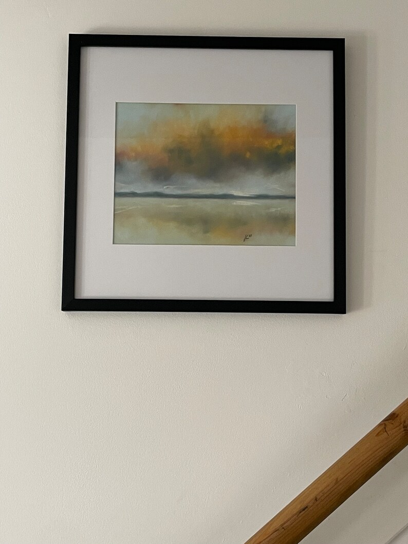 May include: A framed painting of a seascape with a cloudy sky in shades of yellow, orange, and gray. The water is calm and reflects the sky. The painting is in a black frame.