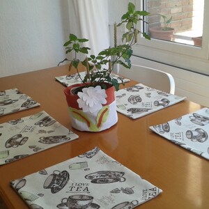 Könnte beinhalten: Ein Set aus fünf rechteckigen Tischsets aus Stoff mit einem wiederkehrenden Muster aus Teetassen und dem Text I LOVE TEA in Braun- und Grüntönen, ergänzt durch einen zylindrischen Übertopf aus Stoff mit einer weißen Blume und grüner Applikation.
