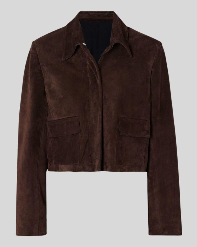 May include: A dark brown suede jacket featuring a collared neckline and two front pockets. The jacket has a cropped length and long sleeves. The color is a rich, dark brown.