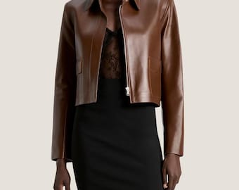 May include: A brown leather jacket with a collared neckline and a zipper closure. The jacket has two front pockets and a cropped length. The model is wearing a black dress underneath.