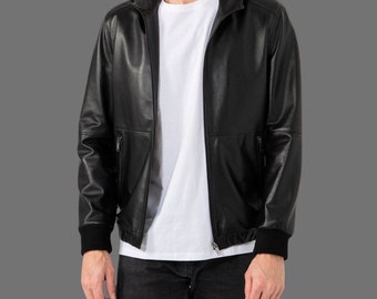 May include: A black leather hooded jacket with a zip-up front, two side pockets, and black ribbed cuffs. The jacket is worn over a white t-shirt and black jeans. The jacket has a classic bomber style.