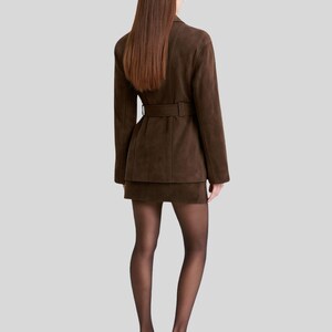 Op de afbeelding: Een bruine su&egrave;de blazer met bijpassende rok. De blazer heeft een riem in de taille. De persoon draagt zwarte doorschijnende panty's en zwarte hakken. De outfit is monochroom.