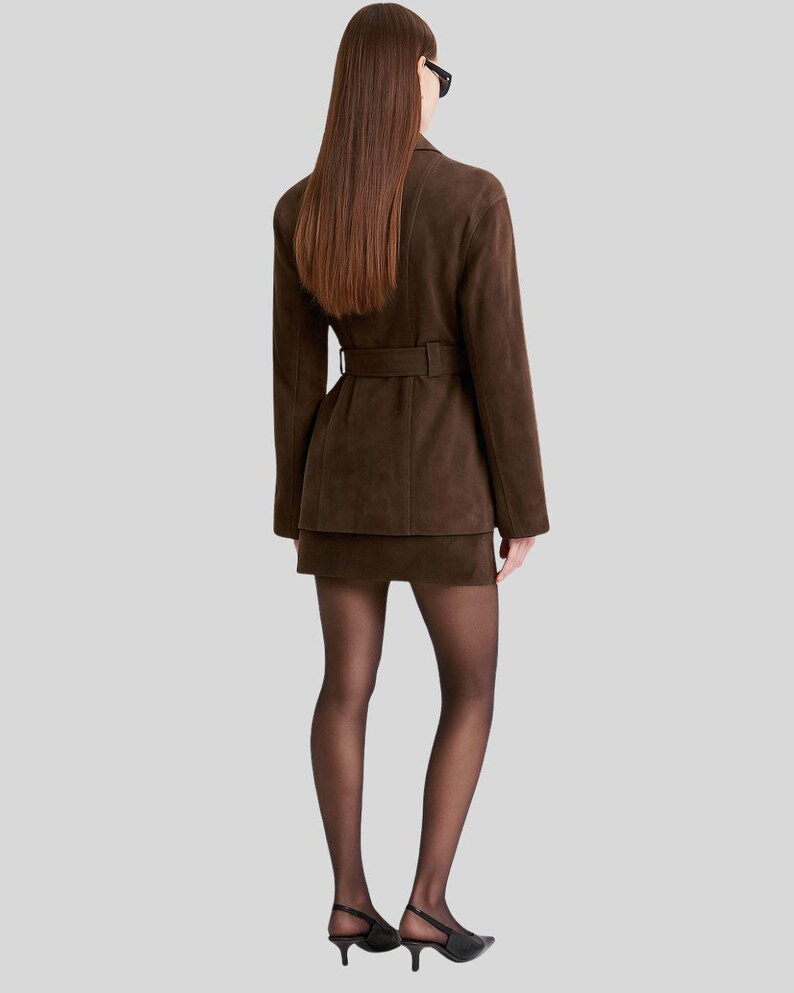 Op de afbeelding: Een bruine su&egrave;de blazer met bijpassende rok. De blazer heeft een riem in de taille. De persoon draagt zwarte doorschijnende panty's en zwarte hakken. De outfit is monochroom.