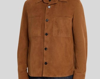 May include: A brown suede jacket with a collared neckline and two front pockets. The jacket has long sleeves and is buttoned up the front with black buttons. The model is wearing dark blue jeans.