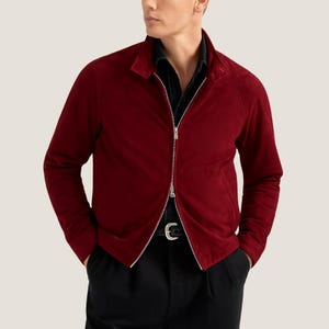 May include: A burgundy suede jacket with a silver zip. The jacket has a collared neckline and long sleeves. The person in the image is wearing a black shirt and black trousers.