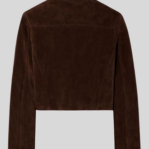 May include: A cropped brown suede jacket with a collared neckline and long sleeves. The back of the jacket is visible, showcasing the tailored seams and a yoke detail. The jacket is a solid, rich brown colour.