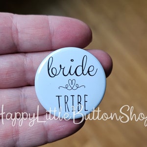 May include: White button with black text that reads "bride tribe" with a small heart symbol above the word "tribe".