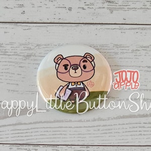 May include: A circular pinback button featuring a cartoon illustration of a brown bear wearing glasses and a yellow bowtie, holding a purple book. The text JOJO APPLE is printed in red, and the text HappyLittleButtonShop is printed in white.