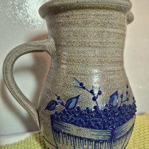 May include: A speckled beige ceramic pitcher with a single handle and a flared rim. The body features a raised blue relief design depicting a basket overflowing with small round berries and leaves, rendered in a deep cobalt blue glaze.
