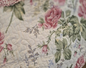 May include: Close-up of a quilted floral pattern. The quilt features large pink roses, green leaves, and small blue flowers on a white background. The edge has a red and white checkered pattern.