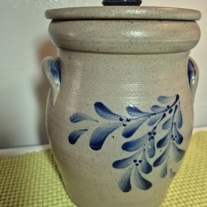 May include: A light gray stoneware crock with a matching lid and two side handles. The exterior features a hand-painted blue floral pattern with small dots. The surface has a speckled texture and a glossy finish, with blue accents on the handles.