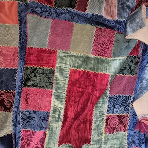 May include: A patchwork quilt with a variety of velvet fabrics in shades of red, green, blue, and gray. The quilt features a blue border with a scalloped edge and gold stitching. The quilt is a cozy home decor item.