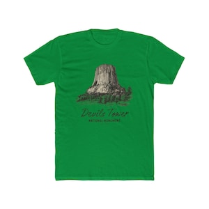 May include: Green t-shirt featuring a graphic of Devils Tower National Monument. The design includes a detailed illustration of the tower and surrounding trees, with the text "Devils Tower" and "National Monument" below the image.