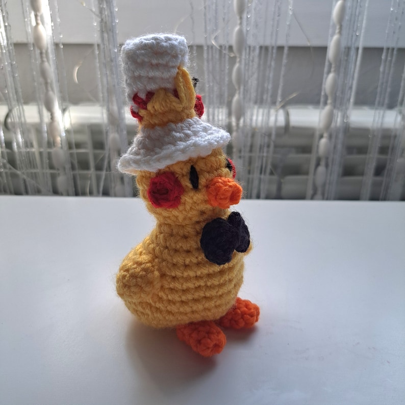 May include: A handmade yellow crochet duckling wearing a white chef's hat and a black bow tie. The duckling has orange feet and a beak, with red accents on its head. This whimsical amigurumi is a charming decorative item.