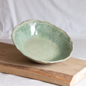 Könnte beinhalten: Handgefertigte Keramikschale mit organischer Form. Die Schale hat eine hellgrüne Glasur mit Farbvariationen. Sie steht auf einem Holzbrett, was ihre handwerkliche Qualität unterstreicht. Durchmesser ca. 20 cm.