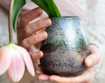May include: A hand holds a small, blue and brown ceramic vase with a rough, textured surface. A pink tulip stem with green leaves is in the vase.