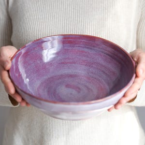 Könnte beinhalten: Eine handgefertigte Keramikschale mit einer glänzenden, wirbelnden lila Glasur. Die Schale hat eine breite, flache Form und einen rötlich-braunen Rand. Das Innere der Schale weist eine Mischung aus lila und weißen Farbtönen auf.