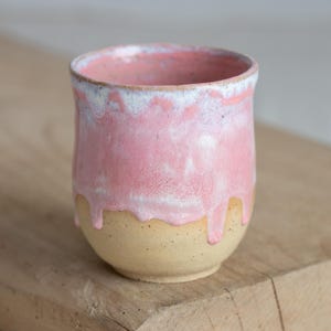 Pode incluir: Uma chávena de cerâmica artesanal com uma cobertura rosa que escorre sobre uma base bege. A borda da chávena tem um detalhe branco. A chávena está sobre uma superfície de madeira.