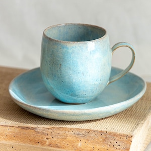 Könnte beinhalten: Ein hellblaues Porzellan-Teetassen-Set. Die Tasse hat eine abgerundete Form und einen Henkel. Die Untertasse ist eine einfache runde Form.