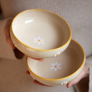 Può includere: Due ciotole in ceramica beige con bordi gialli e disegni di margherite bianche. Le ciotole sono impilate l'una sull'altra.
