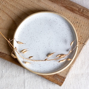 Könnte beinhalten: Ein gesprenkelter weißer Keramikteller mit hellem beigen Rand. Der Teller steht auf einem Holzschneidebrett. Ein getrockneter Pflanzenstiel mit Samenkapseln liegt über dem Teller.