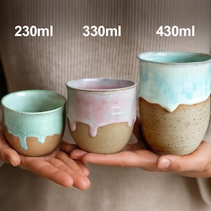 May include: Three handmade ceramic mugs in varying sizes, with text indicating their capacity: 230ml, 330ml, and 430ml. The mugs feature a two-tone design, with a light-colored glaze dripping over a speckled, earthy base. The colors include mint green, pink, and light blue.