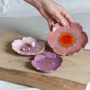 May include: Three ceramic flower-shaped trinket dishes in shades of pink and purple. The largest dish has an orange center. One dish holds a ring and small beads, another holds a necklace. The dishes are on a wooden surface.
