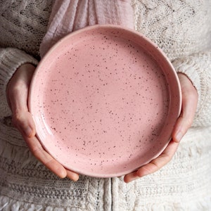 Könnte beinhalten: Ein gesprenkelter rosa Keramikteller. Der Teller ist rund mit einem leicht erhöhten Rand. Die rosa Farbe hat kleine, dunkelbraune Sprenkel. Der Teller wird von einer Person gehalten, die einen weißen Pullover trägt.