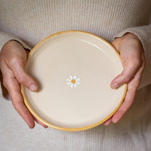 Könnte beinhalten: Ein cremefarbener Keramikteller mit gelbem Rand und einer weißen, in die Mitte gemalten Margerite.